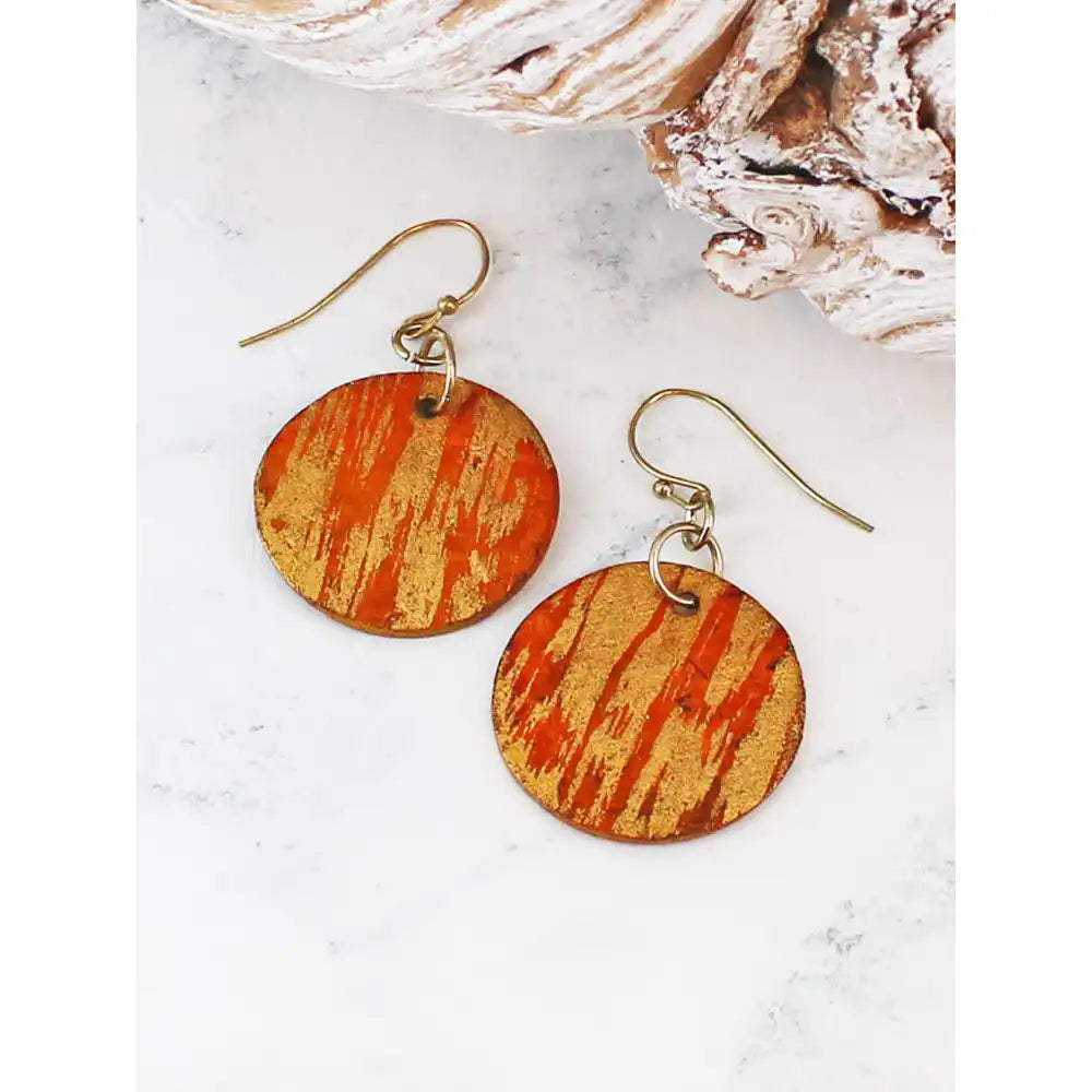 A pair of round drop earrings with a textured orange and gold patterned face and gold-tone fishhook wires, displayed on a white marble surface next to a seashell.