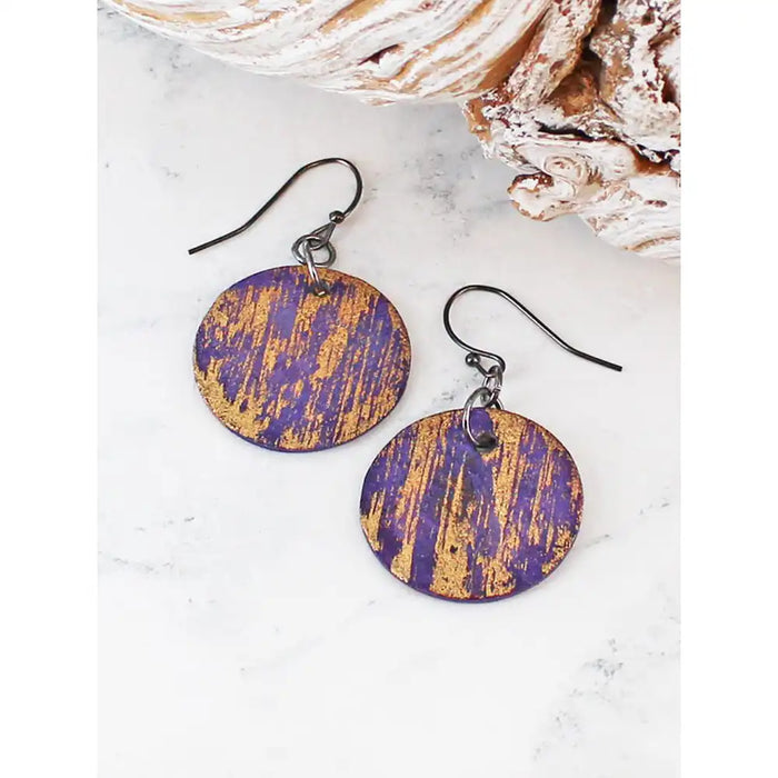 A pair of round drop earrings with a distressed purple and gold pattern, suspended from silver metal fishhook wires, displayed on a white marble surface with a seashell prop.