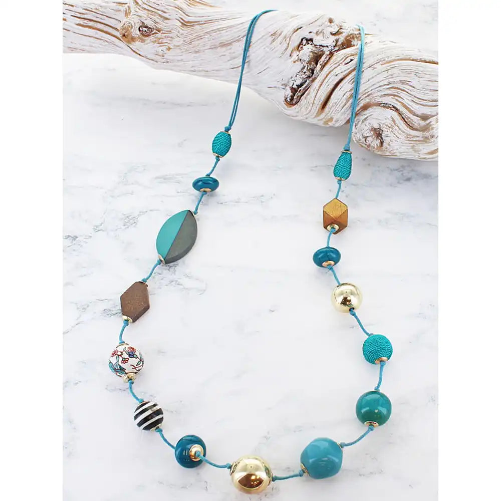 A beaded necklace with turquoise blue cords and multiple colorful beads, displayed on a white marble surface next to a piece of driftwood.
