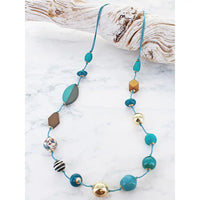 A beaded necklace with turquoise blue cords and multiple colorful beads, displayed on a white marble surface next to a piece of driftwood.
