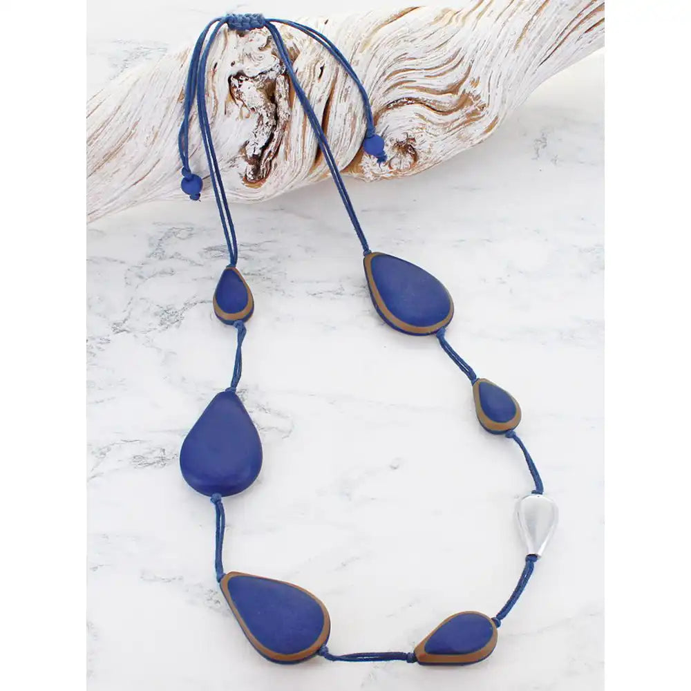 A handmade beaded necklace with blue teardrop-shaped beads outlined in brown, suspended on a blue cord with an adjustable sliding knot, displayed on a white marble surface next to a piece of driftwood.