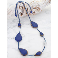 A handmade beaded necklace with blue teardrop-shaped beads outlined in brown, suspended on a blue cord with an adjustable sliding knot, displayed on a white marble surface next to a piece of driftwood.