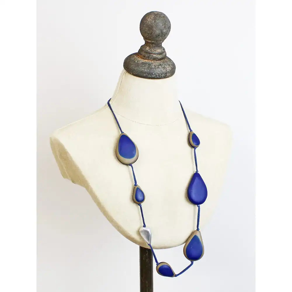 A long statement necklace with large teardrop-shaped blue beads outlined in gold, displayed on a cream-colored fabric mannequin bust against a white background.