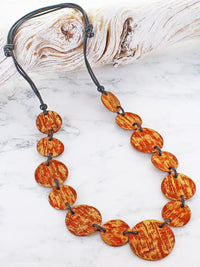 A beaded necklace made of oval shell beads with a textured orange and brown grain pattern, strung on a black cord, displayed on a white marble surface with a piece of driftwood in the background.
