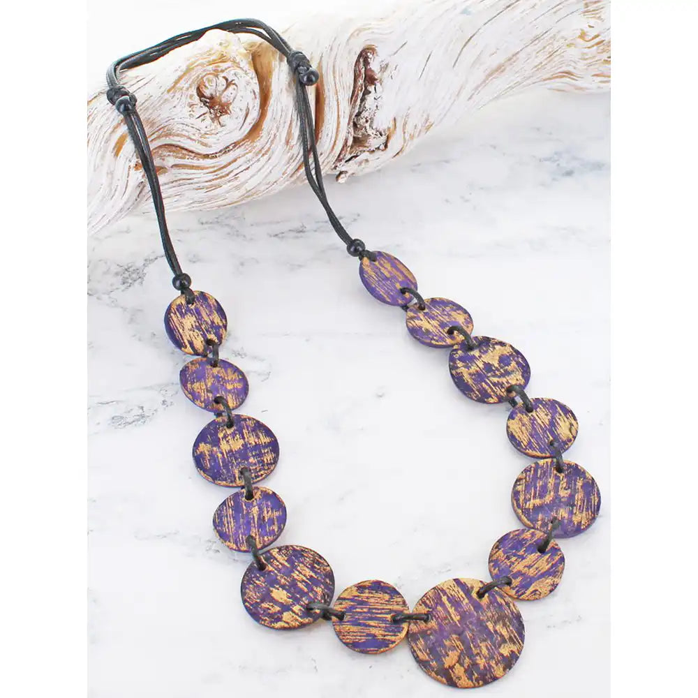 A beaded necklace with oval purple and gold marbled beads on a black cord, displayed on a white marble surface next to a piece of driftwood.