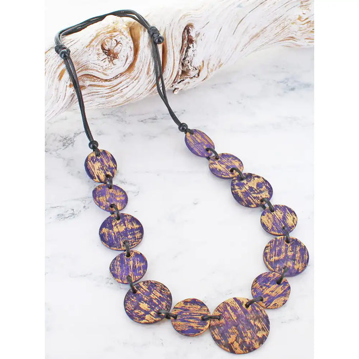A beaded necklace with oval purple and gold marbled beads on a black cord, displayed on a white marble surface next to a piece of driftwood.