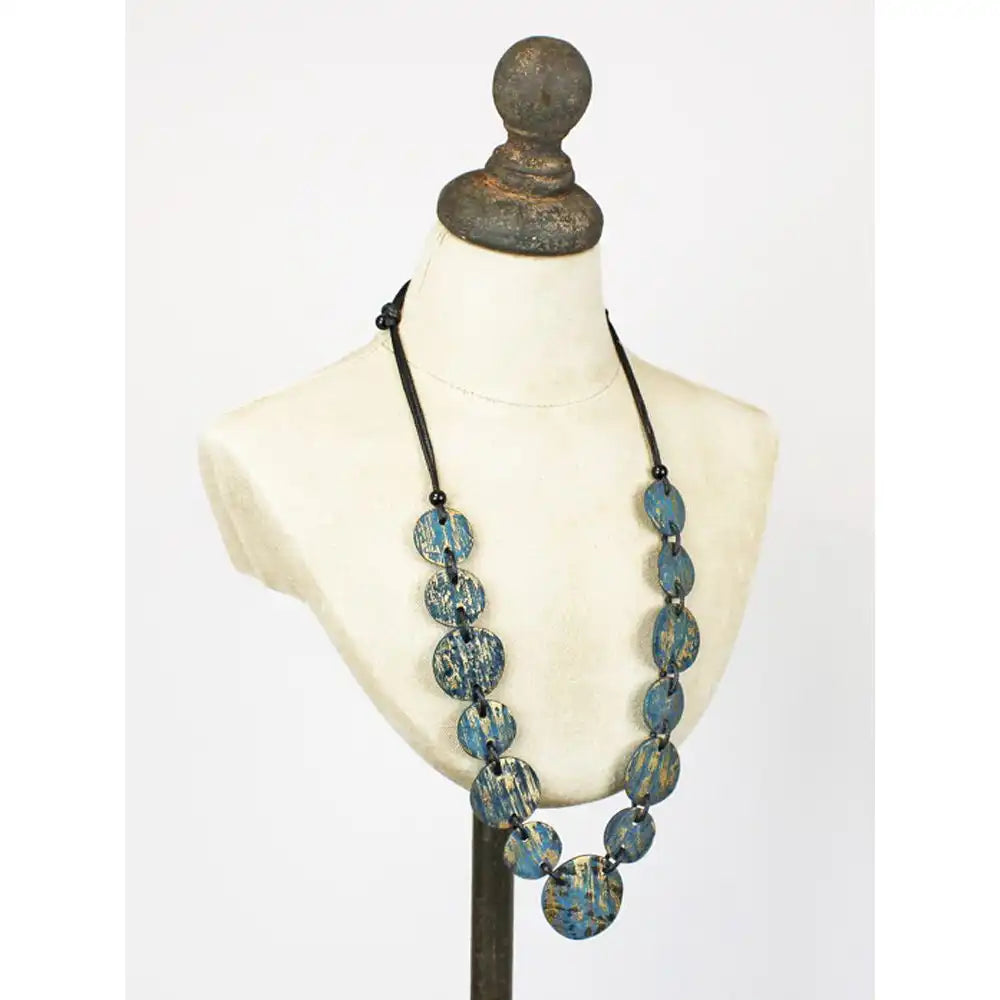 A statement necklace with distressed teal and turquoise wooden beads on a black cord, displayed on a cream-colored mannequin bust.