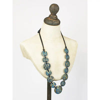 A statement necklace with distressed teal and turquoise wooden beads on a black cord, displayed on a cream-colored mannequin bust.