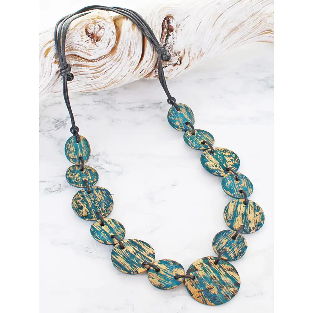 A statement necklace made of oval shell beads with a distressed teal and gold finish, strung on a black cord, displayed on a white marble surface next to a piece of driftwood.