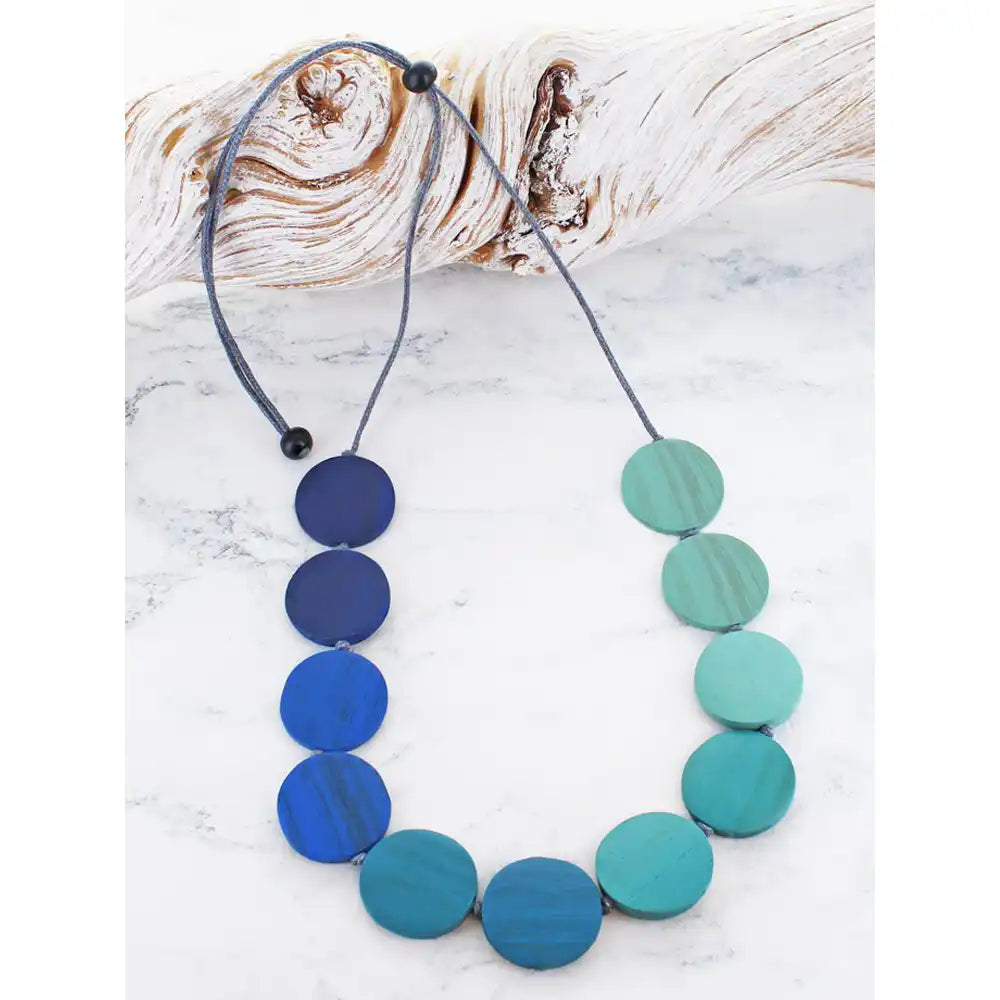 A beaded necklace made of round disc-like beads in shades of blue, suspended on a grEy cord with black toggle beads, displayed on a white marble surface next to a piece of driftwood
