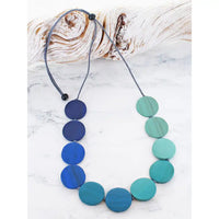 A beaded necklace made of round disc-like beads in shades of blue, suspended on a grEy cord with black toggle beads, displayed on a white marble surface next to a piece of driftwood