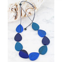 A leaf-shaped beaded necklace in shades of blue, including teal, navy and cobalt, strung on a grey cord with black beads. The necklace is displayed on a white marble surface next to a piece of driftwood.