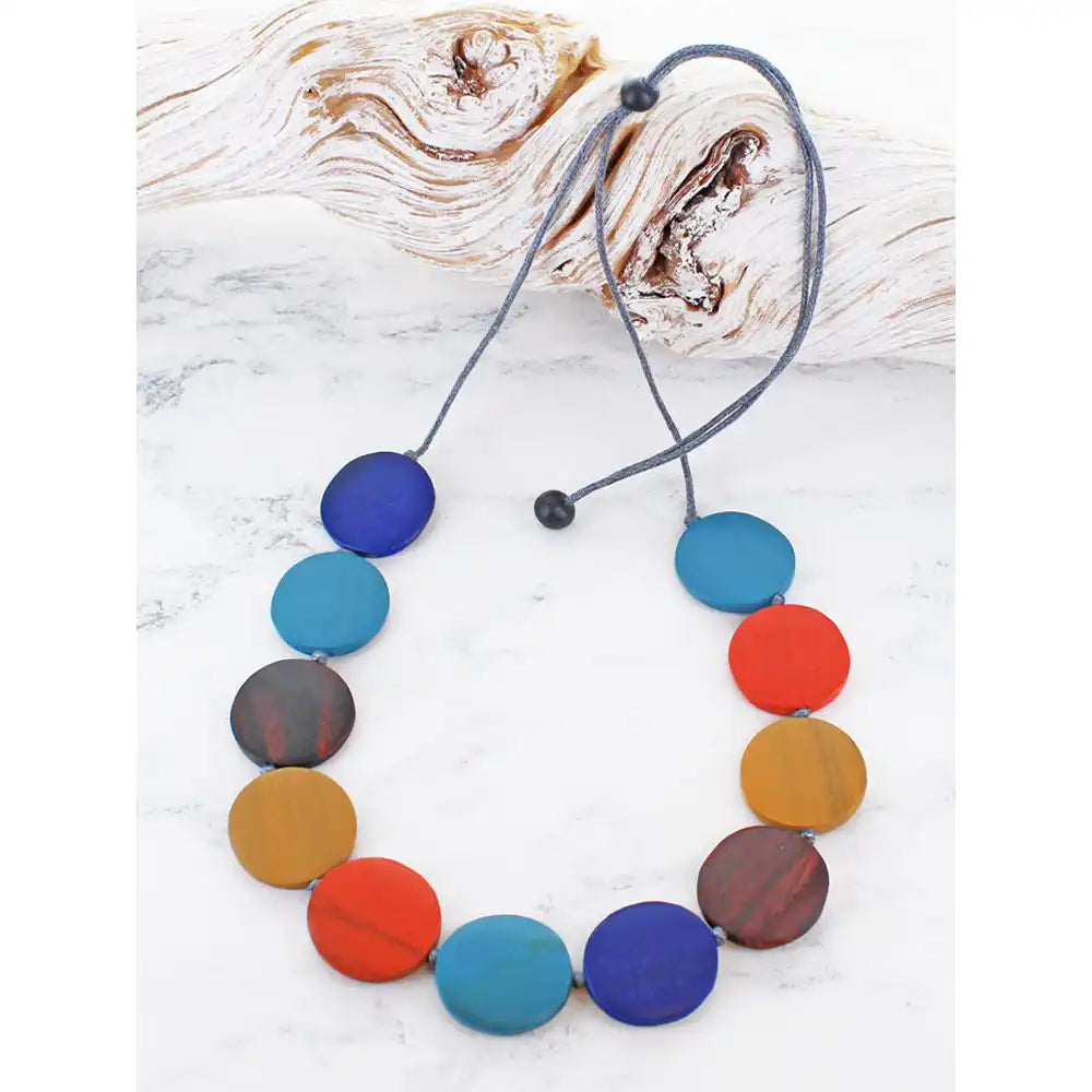 A colorful beaded necklace with round wooden disc beads in shades of blue, teal, red, orange, yellow, and brown, threaded on a grey cord with black toggle beads, displayed on a white marble surface with a piece of driftwood.