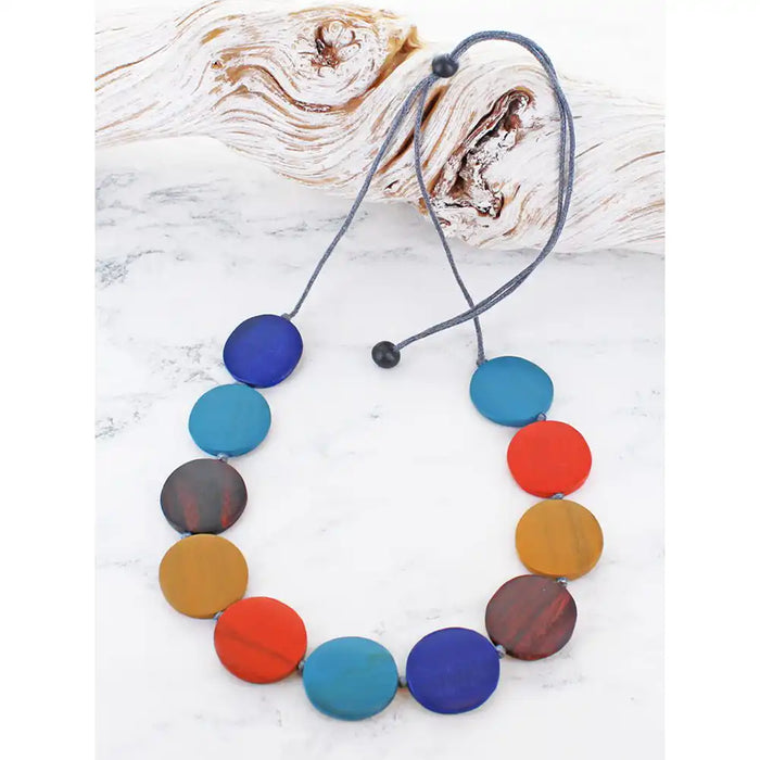A colorful beaded necklace with round wooden disc beads in shades of blue, teal, red, orange, yellow, and brown, threaded on a grey cord with black toggle beads, displayed on a white marble surface with a piece of driftwood.