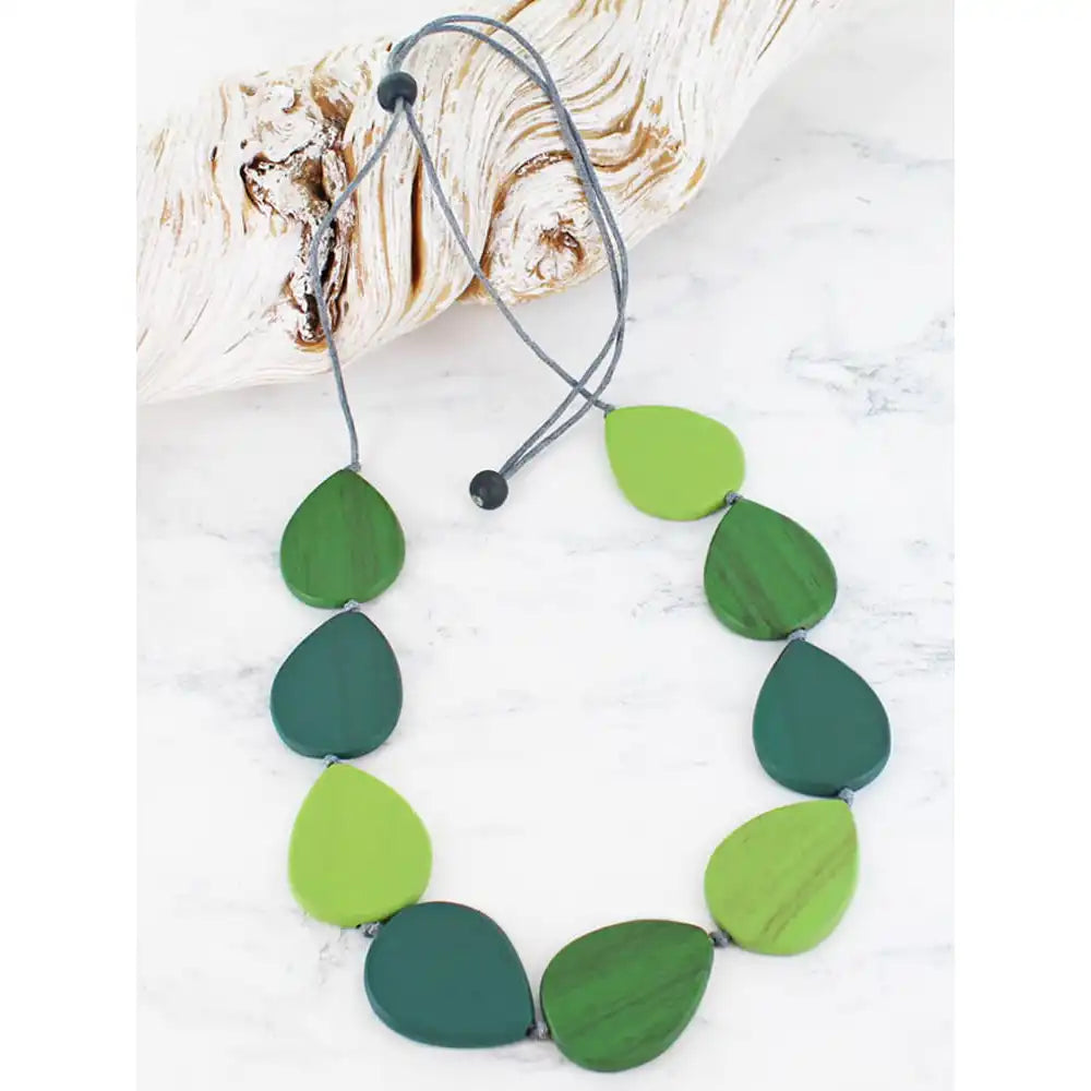 A leaf-shaped beaded necklace in shades of green, including forest green, emerald green, and lime green, strung on a grey cord with black beads. The necklace is displayed on a white marble surface next to a piece of driftwood.
