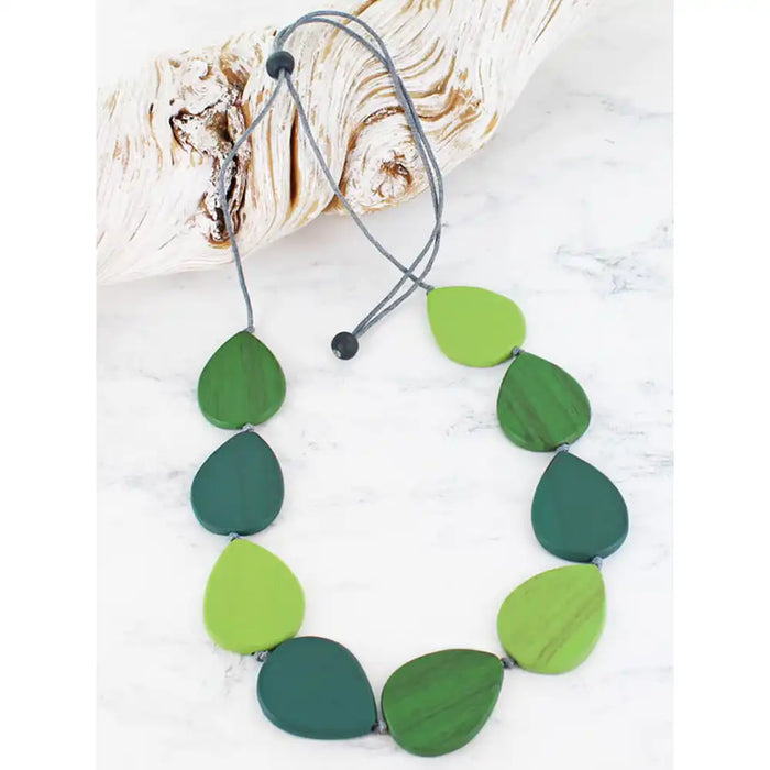 A leaf-shaped beaded necklace in shades of green, including forest green, emerald green, and lime green, strung on a grey cord with black beads. The necklace is displayed on a white marble surface next to a piece of driftwood.