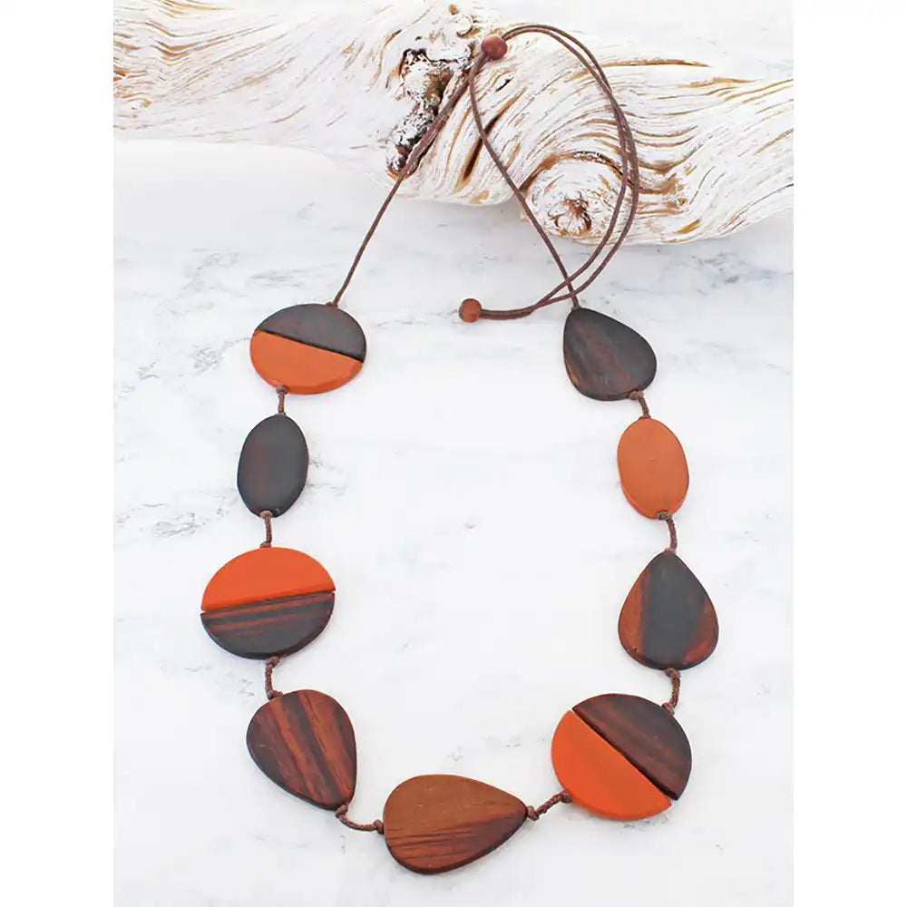 A beaded necklace made of alternating oval and teardrop-shaped wooden beads in dark brown and orange-brown wood, strung on a brown cord with a toggle clasp, displayed on a white marble surface next to a piece of driftwood.