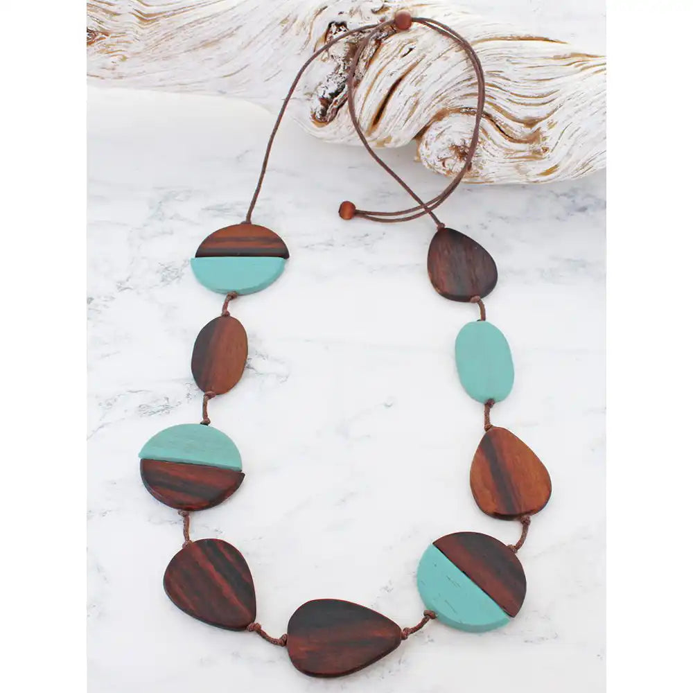 A wooden bead necklace with alternating oval and teardrop-shaped beads in dark brown and turquoise, strung on a brown cord, displayed on a white marble surface with a piece of driftwood.
