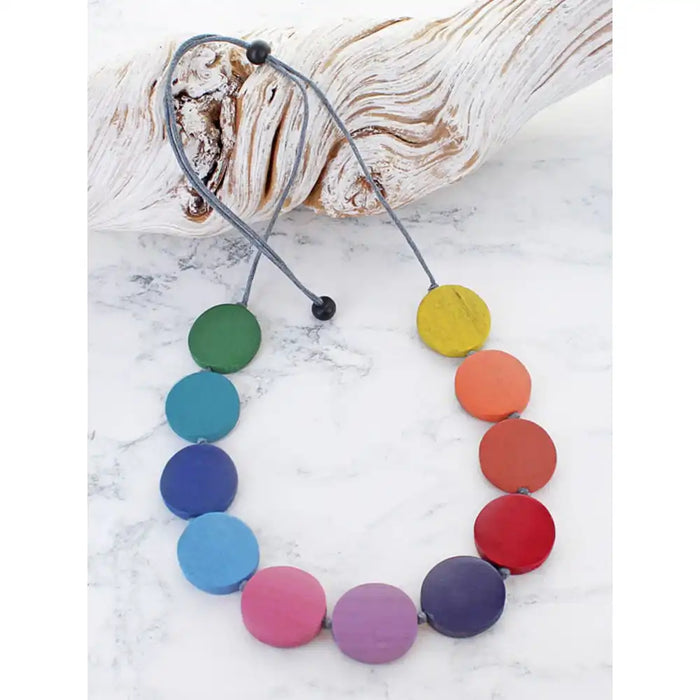 A circular beaded necklace with flat, round wooden discs in a rainbow color sequence, displayed on a white marble surface next to a piece of driftwood.