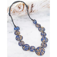 A beaded necklace with oval blue and gold textured beads on a black cord, displayed on a white marble surface with a piece of driftwood in the background.