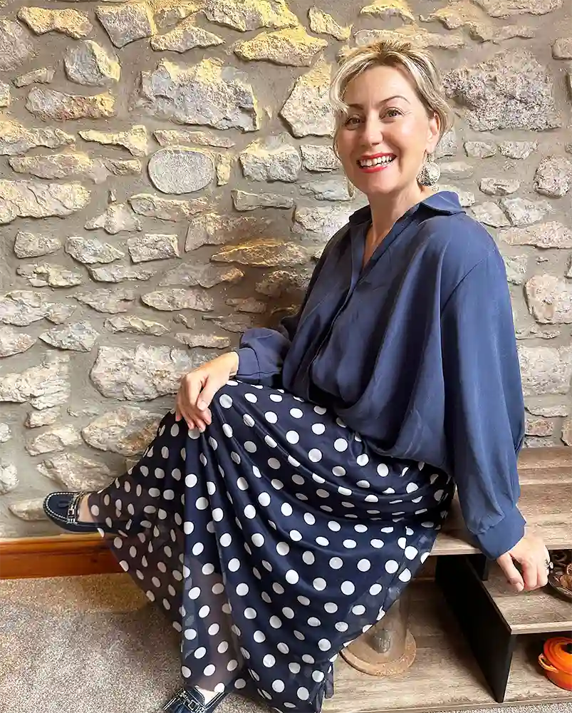 A woman sitting on a wooden bench in front of a rustic stone wall, wearing a navy blue long-sleeve blouse and a navy blue skirt with large white polka dots.