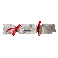 A Christmas-themed gift cracker package wrapped in white paper with a festive print of deer, pinecones, mistletoe, and red berries, tied with red ribbons and accompanied by a small paper tag detailing contents of the cracker.