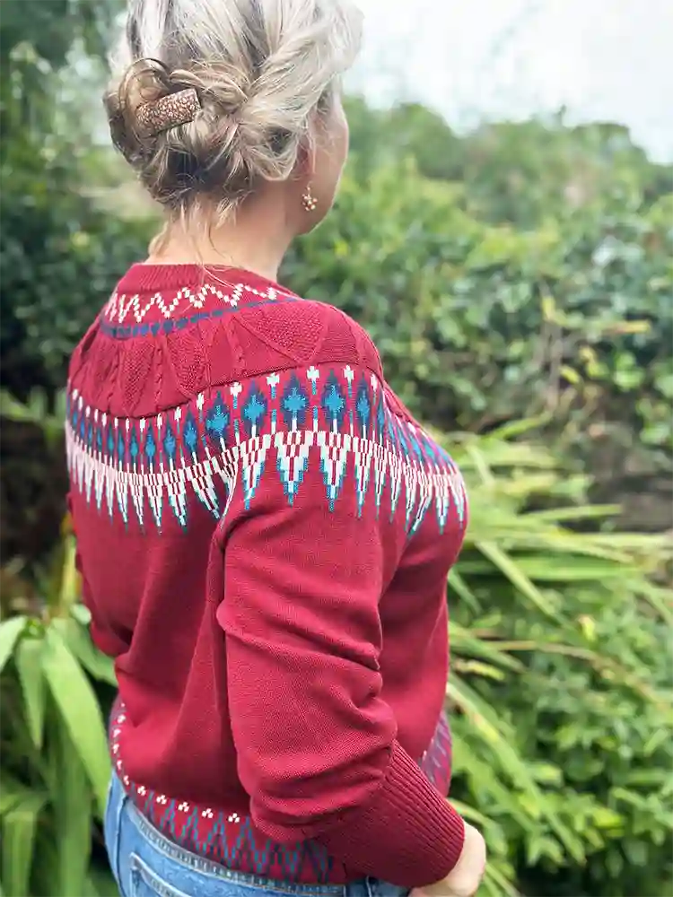 A woman shown from behind wearing a red knit sweater with a blue and white geometric pattern across the shoulders and upper back, standing outdoors in front of green foliage.