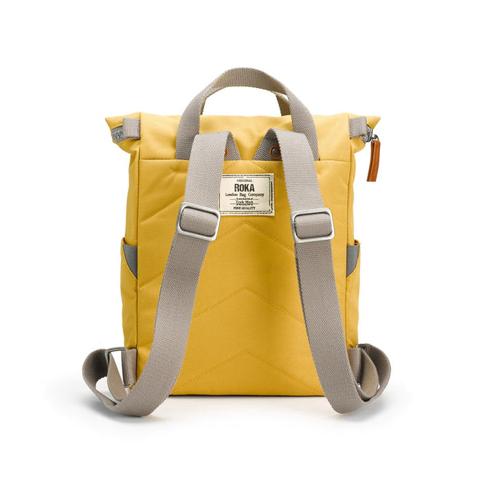Roka flax yellow backpack. Foldover and zip fastening at the top. External zip fastening pocket. Grey handles and back pack straps. Two water bottle pockets. Rear view showing padding and Roka label.